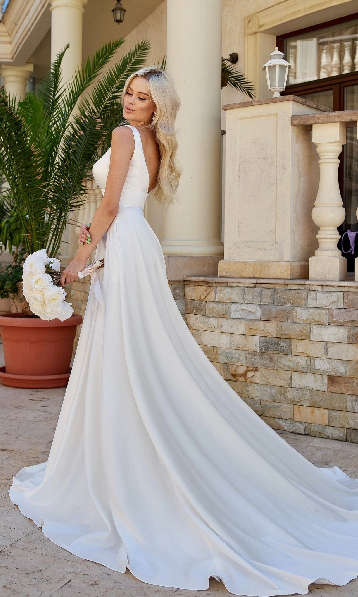 A woman in the Long White Satin Bridal Gown: Tina Holly BA269W stands outdoors on a stone patio, holding a bouquet near a potted palm and a columned building.